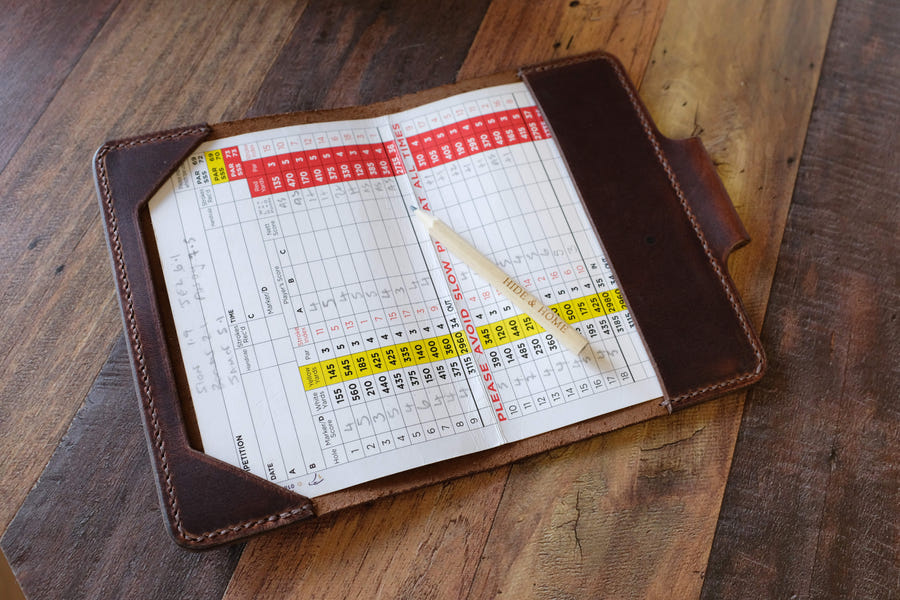 Open brown leather golf scorecard holder with pen loop, holding a scorecard and pencil on a wooden workbench.